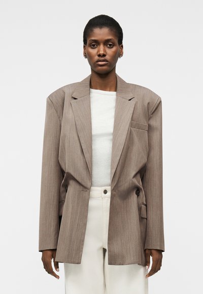 Personne portant un blazer taupe surdimensionné par-dessus un haut blanc et un pantalon crème taille haute, debout devant un fond clair uni.