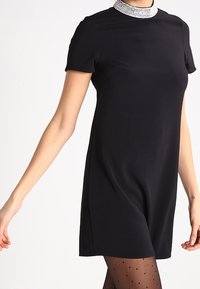 Svart kortärmad klänning med en figurnära silhuett och en glittrande kragsdetalj. Slät tygkvalitet och knälång design, kombinerad med prickiga strumpbyxor.