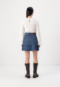 Blouse blanche à manches longues avec des détails texturés, associée à une jupe en denim bleu avec un ourlet volanté et des bottes en cuir marron.