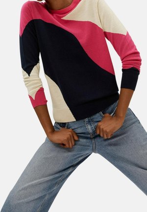 Osoba nosząca sweter z długim rękawem w kolorach różowym, czarnym i beżowym oraz jasnoniebieskie dżinsy, z rękami trzymającymi pas spodni.