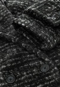 Gros plan d’un manteau texturé en laine noire et grise avec de gros boutons et un revers plié montrant le motif du tissu duveteux.
