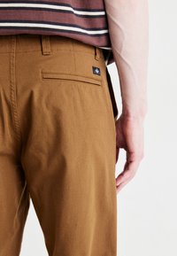Pantalones de algodón marrón con bolsillo trasero, detalle de etiqueta negra y un ajuste relajado, combinados con una camisa de rayas en color burdeos y blanco.