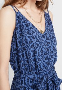 Robe à motifs bleus avec un décolleté en V, des bretelles fines et un détail à nouer à la taille. Le tissu présente un imprimé floral dans des tons de bleu plus foncé.