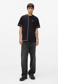 Tricou negru cu mânecă scurtă, cu cusături albe în contrast, croi drept și o etichetă mică cu logo pe piept. Asortat cu blugi gri închis.