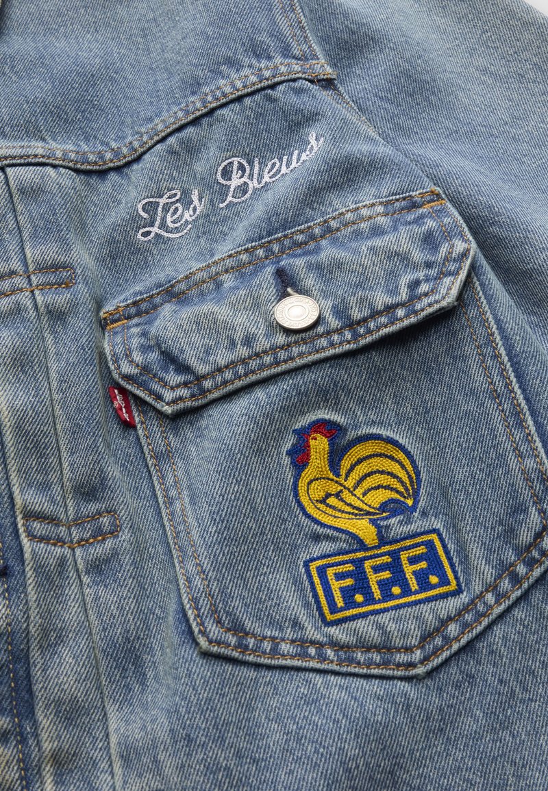 Tasca di giacca in denim azzurro chiaro con gallo giallo ricamato, toppa blu "F.F.F." e testo bianco in corsivo "Les Bleus" sopra la tasca.