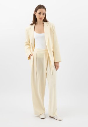 Jonge vrouw staand, gekleed in een crèmekleurig blazer, wijde broek, wit hemdje en witte schoenen tegen een effen witte achtergrond.