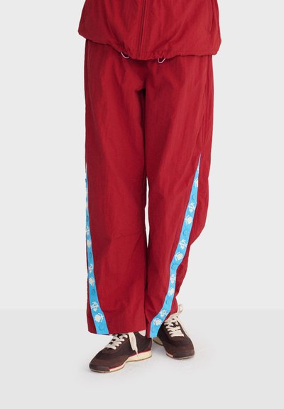 Pantalon large couleur bordeaux avec des bandes latérales bleues à motifs, assorti à des baskets marron et blanches sur fond blanc.
