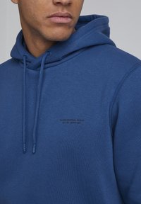 Sudadera de color azul marino hecha de un tejido suave, con un bolsillo canguro en la parte delantera, cordones ajustables y un logo negro impreso en el pecho.