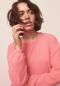 Ružová, štruktúrovaná mikina s rebrovanými manžetami a voľným strihom. Ruka modelky sa dotýka vlasov, na neutrálom pozadí.