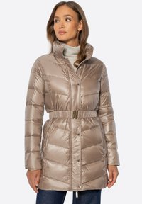 Mujer con una chaqueta acolchada beige brillante con cinturón sobre un suéter de cuello alto claro, de pie frente a un fondo liso y claro.