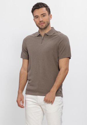 Man die een bruin shirt met korte mouwen en een ritskraag draagt en een witte broek, staand tegen een effen lichte achtergrond.