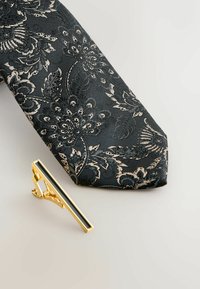 Zwart stropdas met een bloemachtig patroon in zilver- en gouden tinten; vergezeld door een gouden dasclip met een rechthoekige zwarte acccent.