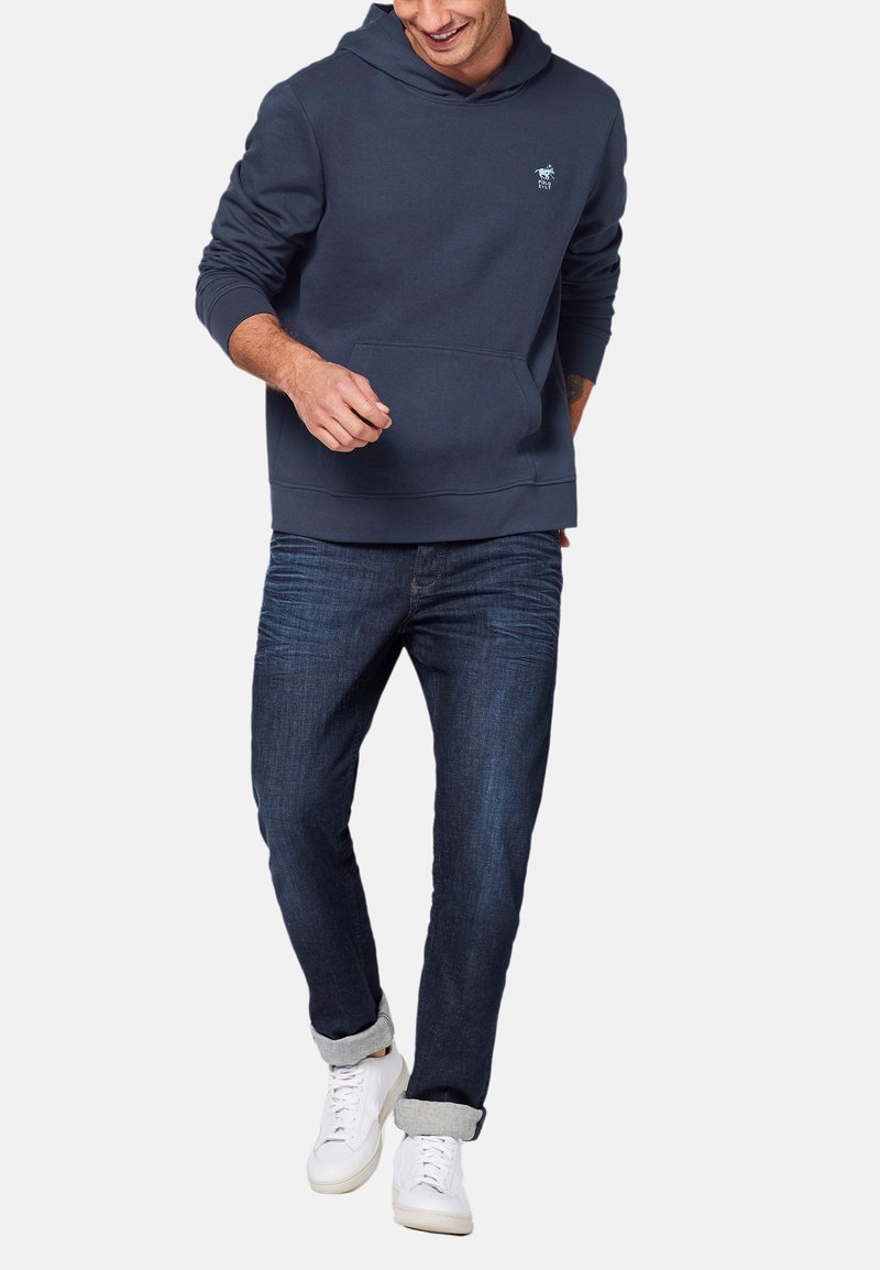 Mørkeblå hætte sweatshirt med en frontlomme og broderet logo, parret med mørke denim jeans og hvide sneakers.