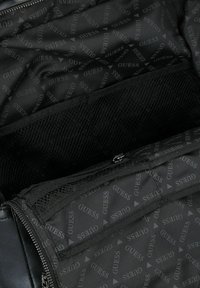 Interno di borsa con rifiniture nere caratterizzato da un motivo a logo "GUESS" ripetuto, tasca in rete e scomparto con zip, con materiale liscio e dettagli testurizzati.