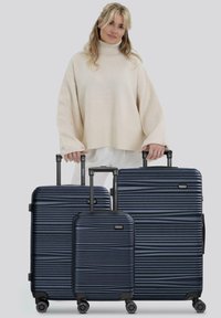 Femme en pull beige tenant les poignées de trois valises rigides bleu marine avec des poignées télescopiques déployées.