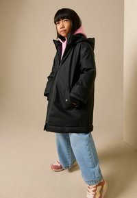 Juoda neperšlampama striukė su gaubtu, derinama su rožine džemperiu su gobtuvu, šviesiai mėlynomis plačiomis kelnėmis ir rožiniais sportbačiais su baltais akcentais.