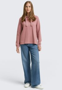 Sudadera color rosado con cordones, combinada con jeans de pierna ancha azules y zapatillas blancas. Textura suave, corte relajado, diseño minimalista.