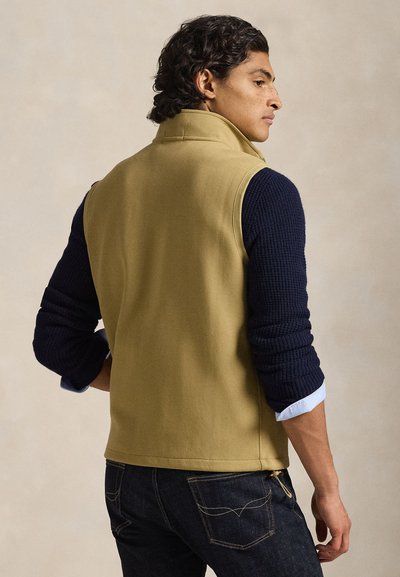 Khakinvärinen hihaton liivi, jossa on resoritaskuiset tummansiniset hihat, korkea kaulus ja sivusaumat. Päällä sininen kauluspaita ja tummansiniset farkut.