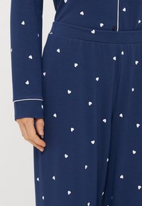 Ναυτική πιτζάμα με λευκό μοτίβο καρδιάς, διαθέτει μακριά μανίκια και κουμπί στο κλείσιμο. Λιγνό ύφασμα με λευκή επένδυση στις μανσέτες.