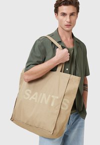 Beige Baumwolltasche mit "SAINTS" in großen Buchstaben bedruckt. Mit zwei langen Tragegriffen. Glatte Textur, quadratische Form.