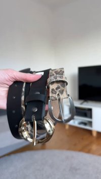 Deux ceintures sont tenues dans une main : une en cuir noir avec des accessoires dorés, l'autre beige à motifs avec des taches de léopard noires et une boucle argentée.