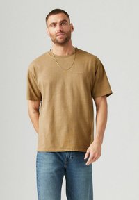 Braunes Baumwoll-T-Shirt mit kurzen Ärmeln und rundem Halsausschnitt. Mit gesticktem Logo auf der Vorderseite und einem lockeren Schnitt. Getragen mit blauen Jeans.