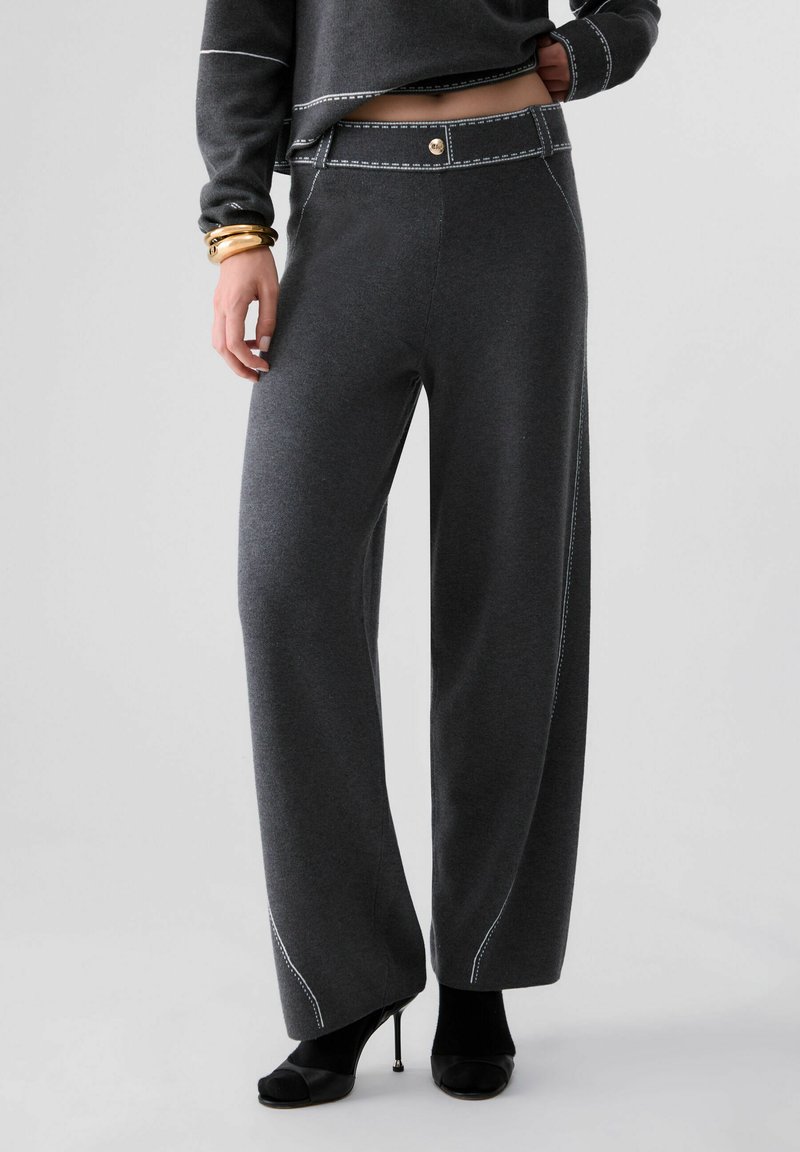 Femme portant un pantalon large gris foncé avec des détails de couture blancs, des chaussures noires à talons ouverts et un top court à manches longues.