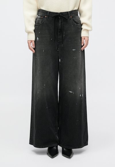 Pantaloni in denim neri, ampi, con vita alta e chiusura a lacci, decorati con macchie di vernice e dettagli di cucitura in contrasto.