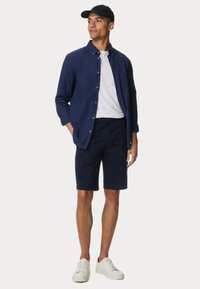 Camicia blu a bottoni sopra una maglietta bianca, abbinata a pantaloni corti blu e sneakers bianche. Il cappellino abbina il colore della camicia.