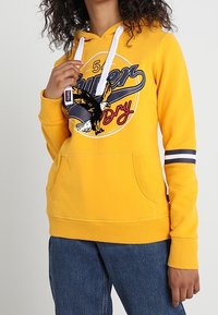 Persona que lleva una sudadera amarilla con un gráfico de un águila y el texto "Super Dry", cordones blancos, detalle a rayas en la manga y jeans azules.