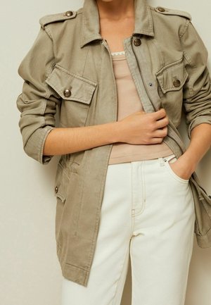 Person trägt beige Utility-Jacke über einem hellrosa Tanktop und weißen Hosen, mit einer Hand in der Tasche und der anderen hält das Revers der Jacke.