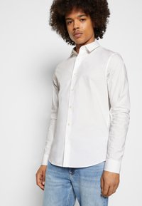 Camicia bianca a maniche lunghe con bottoni, in cotone liscio, colletto classico e vestibilità slim, abbinata a jeans azzurri chiari.