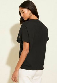 Schwarzes Kurzarmoberteil mit strukturiertem Design, runder Ausschnitt und einem Logoakzent am Ärmel. Kombiniert mit weißen Hosen.