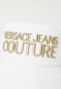 Chapéu de balde branco com bordado em alto-relevo dourado a dizer "VERSACE JEANS COUTURE" na frente. Tecido texturizado com um acabamento suave.