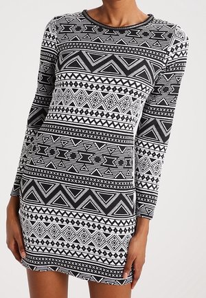 Femme portant une robe à manches longues, noire et blanche avec des motifs tribaux géométriques et un col rond.