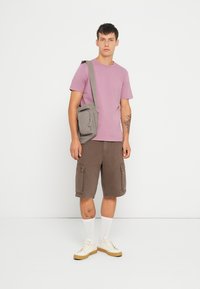Paarse katoenen t-shirt, bruine cargoshorts met zijzakken, grijze schoudertas, witte sokken en beige sneakers. Simpele casual outfit.