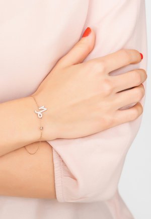 Rose gold bracelet featuring a stylized "M" adorned with clear crystals, linked by a delicate chain with a small drop accent.