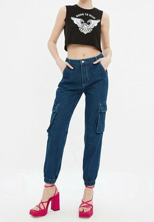 Frau trägt ein schwarzes Crop-Top mit einem Adler-Design und dem Schriftzug "Born to Ride", blaue Cargo-Jeans und leuchtend pinke High-Heel-Sandalen, Hände in den Taschen.