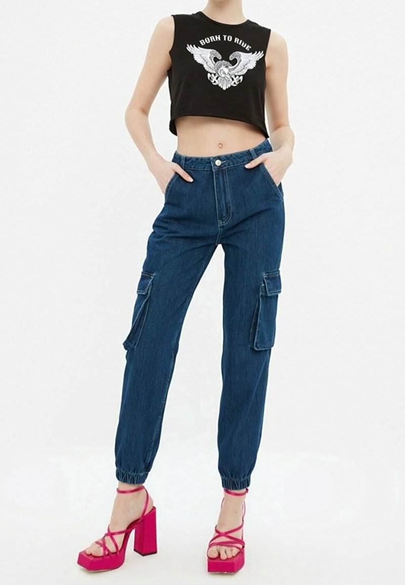 Femme portant un crop top noir avec un graphique d'aigle « Born to Ride », un jean cargo bleu et des sandales à talons hauts rose vif, les mains dans les poches.