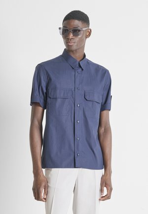 Hombre con camisa azul oscuro de manga corta con botones y dos bolsillos en el pecho, pantalones blancos, mirando hacia adelante con gafas de sol.