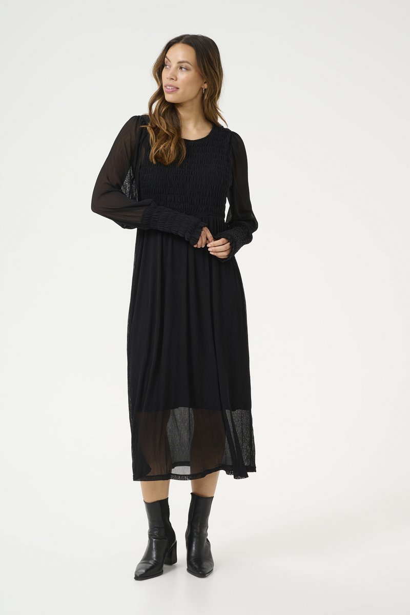 Svart långärmad klänning med smockad bodice och flödande kjol. Genomskinligt underlager. Buren med svarta ankelboots. Texturen verkar lätt.