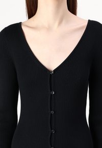 Top en tricot côtelé noir avec un décolleté en V et une rangée de boutons noirs sur le devant, manches longues et coupe ajustée.