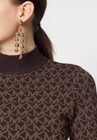 Haut marron à manches longues avec un motif texturé et un col côtelé, associé à des boucles d'oreilles dorées audacieuses ornées de plusieurs accents sphériques.
