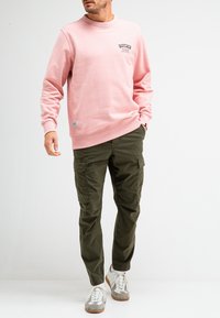 Lichtroze sweatshirt met een ronde hals en logo op de borst, gecombineerd met olijfgroene cargo-broeken en grijze sneakers met witte accenten.