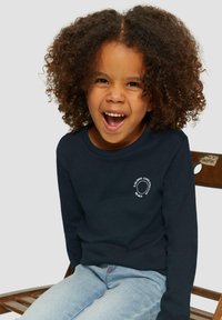 Jong kind met krullend haar, gekleed in een donkerblauwe longsleevenshirt en lichtblauwe spijkerbroek, zittend op een houten stoel en glimlachend met open mond.