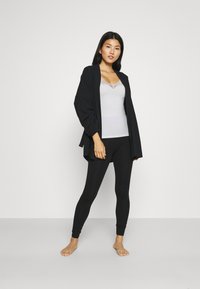 Cardigan noir à manches larges, débardeur gris avec de la dentelle au niveau du col, et leggings noirs. Porté pieds nus sur un fond neutre.