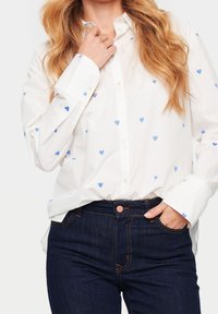 Camicia bianca con motivi a cuore blu, dotata di colletto e maniche lunghe, abbinata a jeans blu scuro e un dettaglio con tasca frontale.