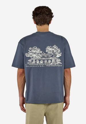 Mann iført en blå t-skjorte med et hvitt trykk av et hus og trær og teksten "Moments to Memories" på baksiden.