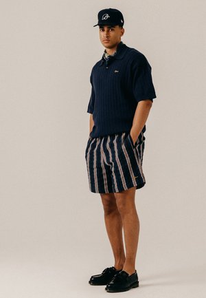 Ung mand iført en mørkeblå kasket, mørkeblå kortærmet strikket polo, stribede mørkeblå og beige shorts og sorte loafers, stående med hænderne i lommerne.