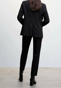 Mustat räätälöidyt puku takki ja housut nähtynä takaa, sileä kangas, tyylikäs muotoilu, istuva leikkaus, yhdistetty kiiltävien mustien nilkkasaappaiden kanssa.
